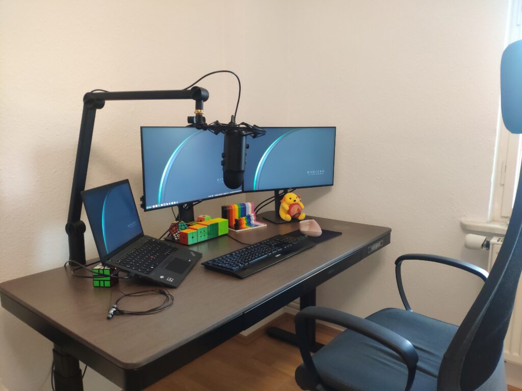 Mein Schreibtisch mit zwei Monitoren, Laptop, Podcast-Mikrofon, Rubiks Cubes, Wapuu-Plüschtier und pinker vertikaler Maus.